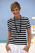 A young blonde woman with short hair on a beach wearing a black-and-white striped top and white summer trousers