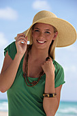 A young blonde woman on a beach wearing a green top and a beige hat