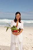 A young brunette woman on a beach wearing a white summer dress and holding a bowl of vegetables