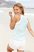 A young blonde woman on a beach wearing a light-blue top and white summer shorts