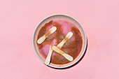 Pink and brown dissolved popsicles with wooden sticks lying in pastel bowl on pink background