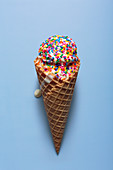 A melting ice cream with colourful sugar sprinkles in a waffle cone, in front of a blue background