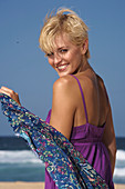 Blonde, kurzhaarige Frau im lila Top mit Strandtuch am Strand