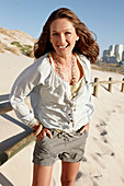 A brunette woman on a sandy beach wearing a light blouse and shorts