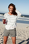 A brunette woman on a sandy beach with a drink wearing a striped top and shorts