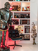 Suit of armour and leather chair in front of shelves holding eclectic collection of items