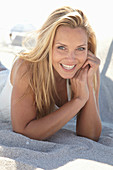 A mature blonde woman lying on a sandy beach wearing a white top