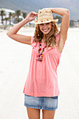 Junge blonde Frau im rosa Top, kurzem Jeansrock und beigem Hut am Strand