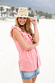 A young blonde woman on a beach wearing a pink top,  a short denim skirt and a beige hat