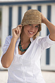 A young blonde woman on a beach wearing a white blouse and a brown hat