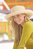 A young blonde woman on a beach wearing an olive top and a beige summer hat