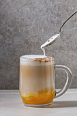 Glass of pumpkin layered spice latte with pumpkin puree, milk foam flowing from spoon and cinnamon