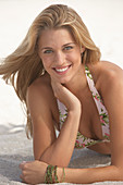 A young blonde woman wearing a patterned bikini lying on a sandy beach