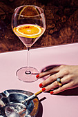 A drink with a blood orange slice, a woman's hand with red fingernails and an ashtray with a cigarette on a bar