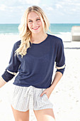 A young blonde woman on a beach wearing a long-sleeved blue shirt and shorts