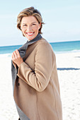 A mature woman with short blonde hair on a beach wearing a grey-brown coat