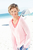 A mature woman with short blonde hair on a beach wearing a pink and white top and colourful trousers