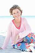 A mature woman with short blonde hair on a beach wearing a pink and white top and colourful trousers