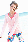 A mature woman with short blonde hair on a beach wearing a pink and white top and colourful trousers