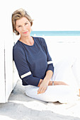 A mature woman with short blonde hair on a beach wearing a blue shirt and white trousers