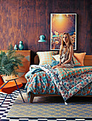 Young woman on double bed with colorful bedding in front of wooden wall