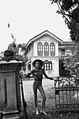 Junge Frau mit Hut, Hose und Bluse steht vor Haus