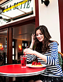 A young brunette woman wearing a striped shirt sitting in a cafe with a slice of cake and a drink