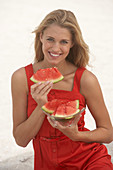 A young blonde woman wearing a red summer dress holding slices of watermelon