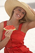 A young blonde woman wearing a red summer dress and a summer hat holding a slice of watermelon