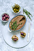 Liver pate in ceramic form, decorated with fresh thyme and two baguette sandwiches with pate, capers, olives on a marble stand