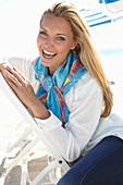 Blonde Frau mit buntem Halstuch und weißer Jacke am Strand