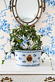 Ivy planted in ceramic pot on vintage chest of drawers below mirror on blue-and-white floral wallpaper