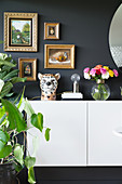 Ceramic leopard's head and vase of flowers on sideboard below gilt-framed pictures on dark wall
