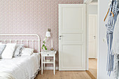 Bedside table next to white metal bed with crocheted blanket in bedroom with floral wallpaper