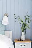 Bedside table next to double bed and wall lamp in bedroom with pale blue wall