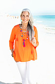 A mature woman with white hair on a beach wearing an orange tunic and white summer trousers