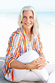 Reife Frau mit weißen Haaren in gestreiftem Hemd und weißer Sommerhose am Strand