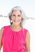 A mature woman with white hair on a beach wearing a pink top