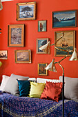 Gemäldesammlung mit Meeresmotiven an roter Wand über Tagesbett mit Kissen, im Vordergrund Stehlampe aus den 1940er Jahren