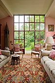 Couch and antique armchairs in front of floor-to-ceiling glass doors leading to garden in living room in shades of pink