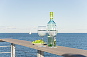 Bottle of wine, glasses and grapes on terrace railing with sea view