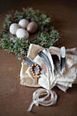 Federn und Kreuz mit Holzperlen auf weißem Tuch, im Hintergrund besprenkelte Eier auf Zinnteller im Osternest