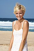 A blonde woman with short hair on a beach wearing a white dress