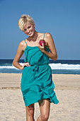 A blonde woman with short hair on a beach holding a pitahaya wearing a turquoise dress