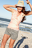 A brunette woman by the sea wearing a hat, an apricot blouse and shorts