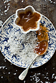 A jar of caramel and a spoon, on a plate with shredded coconut