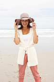 A brunette woman by the sea wearing a short-sleeved cardigan and a hat