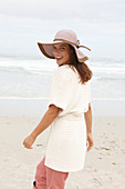 A brunette woman by the sea wearing a short-sleeved cardigan and a hat