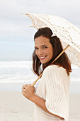 A brunette woman by the sea wearing a short-sleeved cardigan and holding a parasol