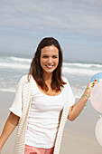 A brunette woman by the sea wearing a short-sleeved cardigan and holding balloons